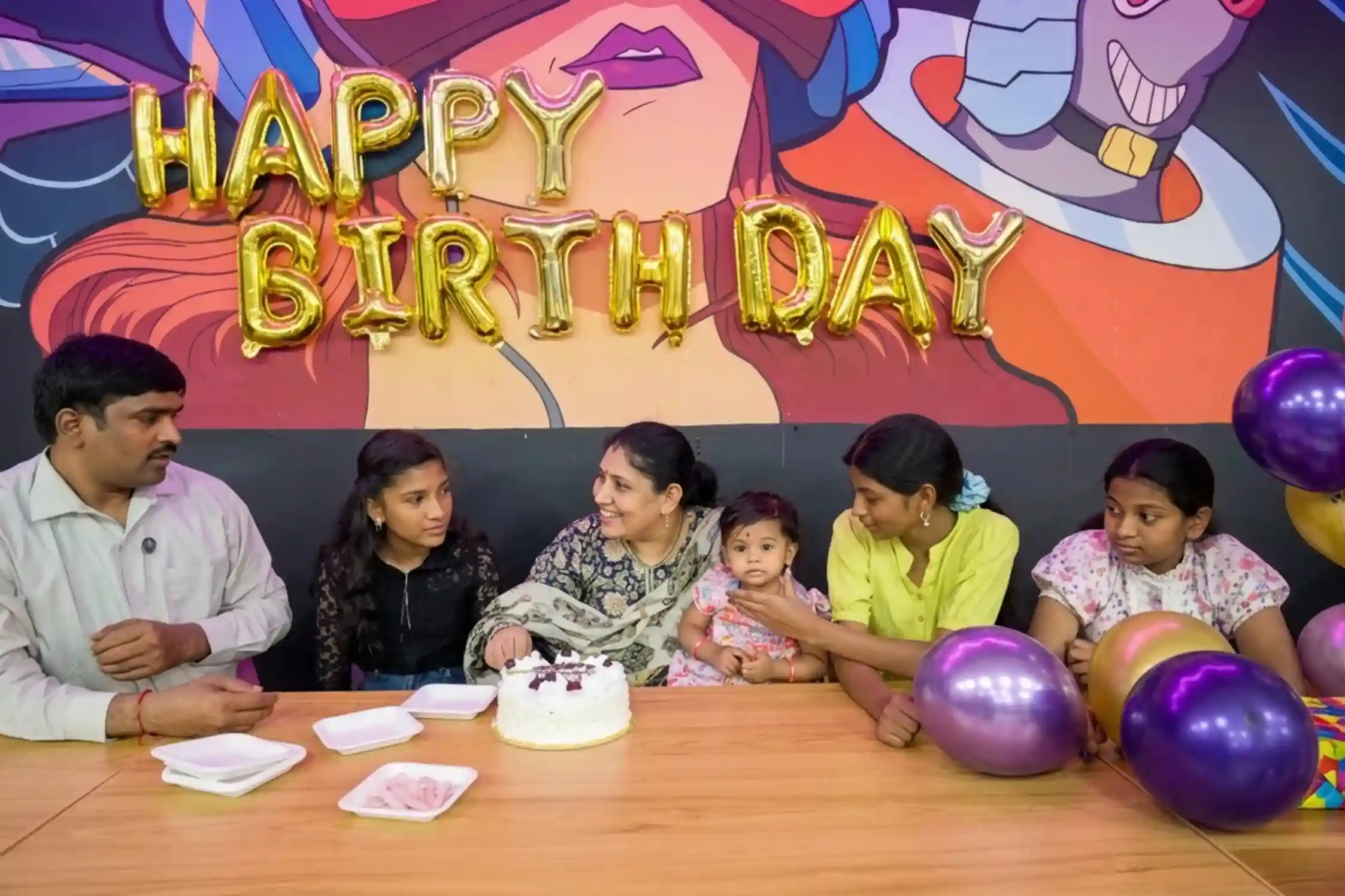 Family gathered around a table with a toddler on grandmother's lap, cake, balloons, and HAPPY BIRTHDAY foil letters
