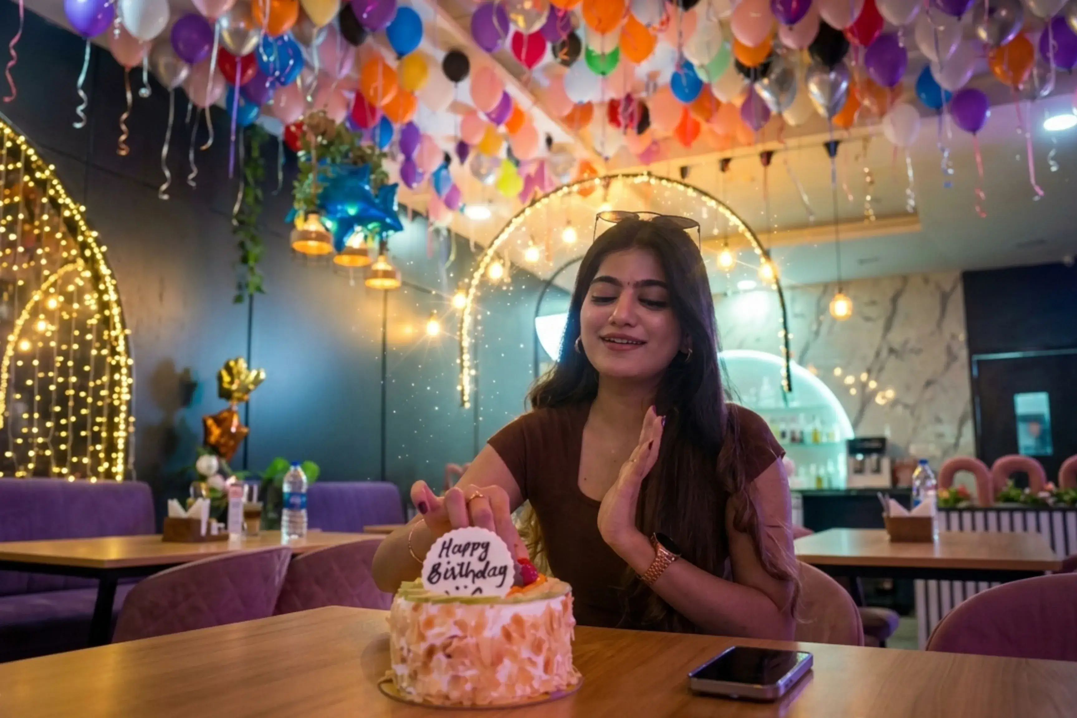 Young woman smiling at her Happy Birthday cake with fairy lights and hundreds of balloons overhead