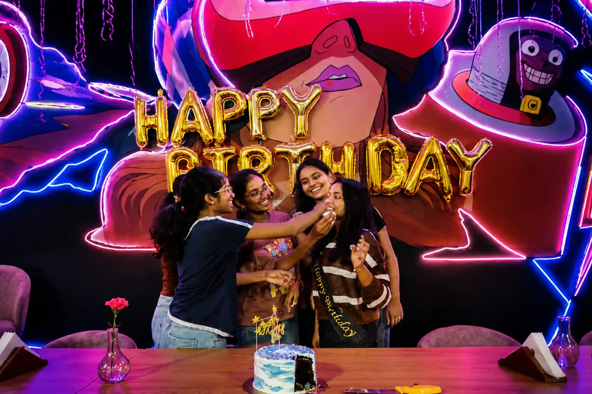 Four teen friends laughing and feeding birthday cake to each other with neon HAPPY BIRTHDAY backdrop